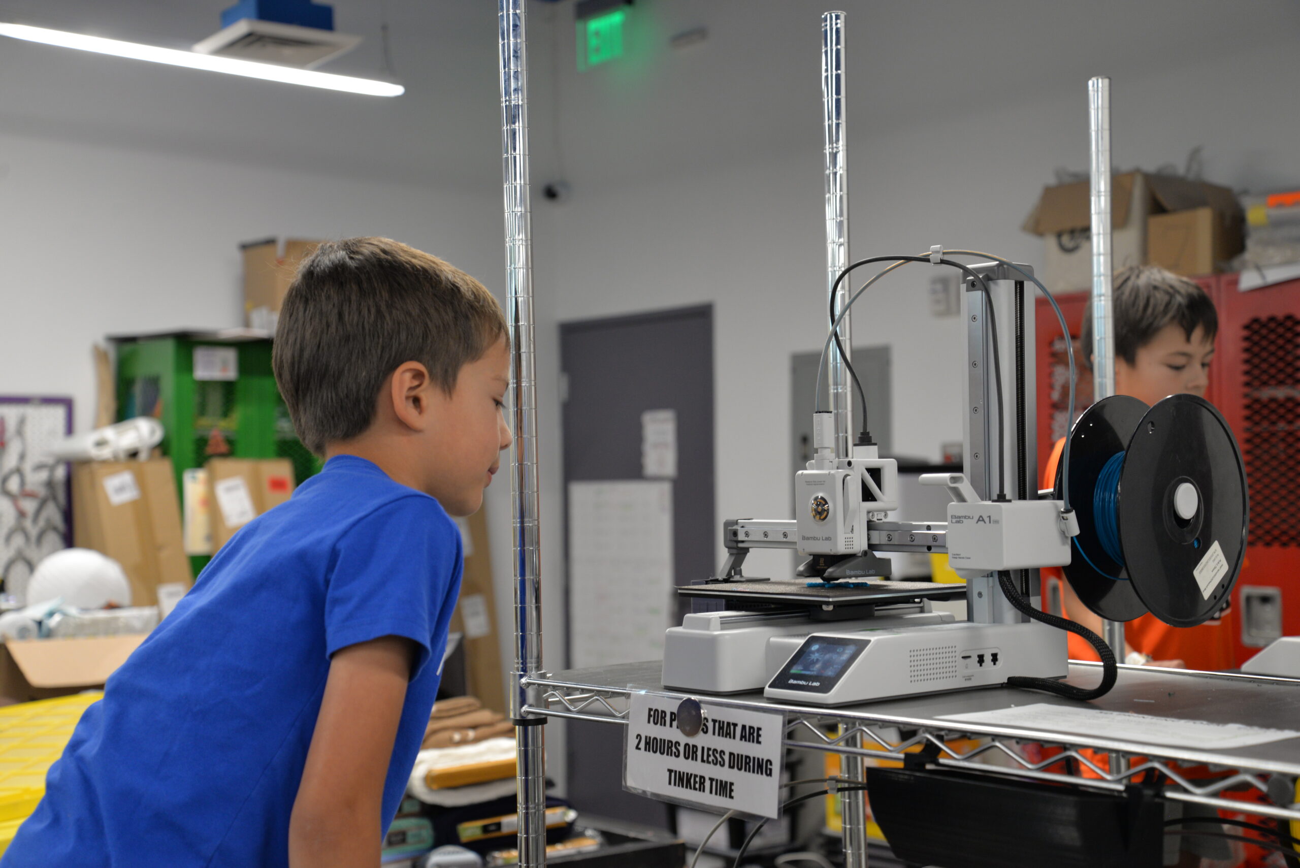 A boy in a blue shirt watches a 3D printer in a classroom or lab setting.