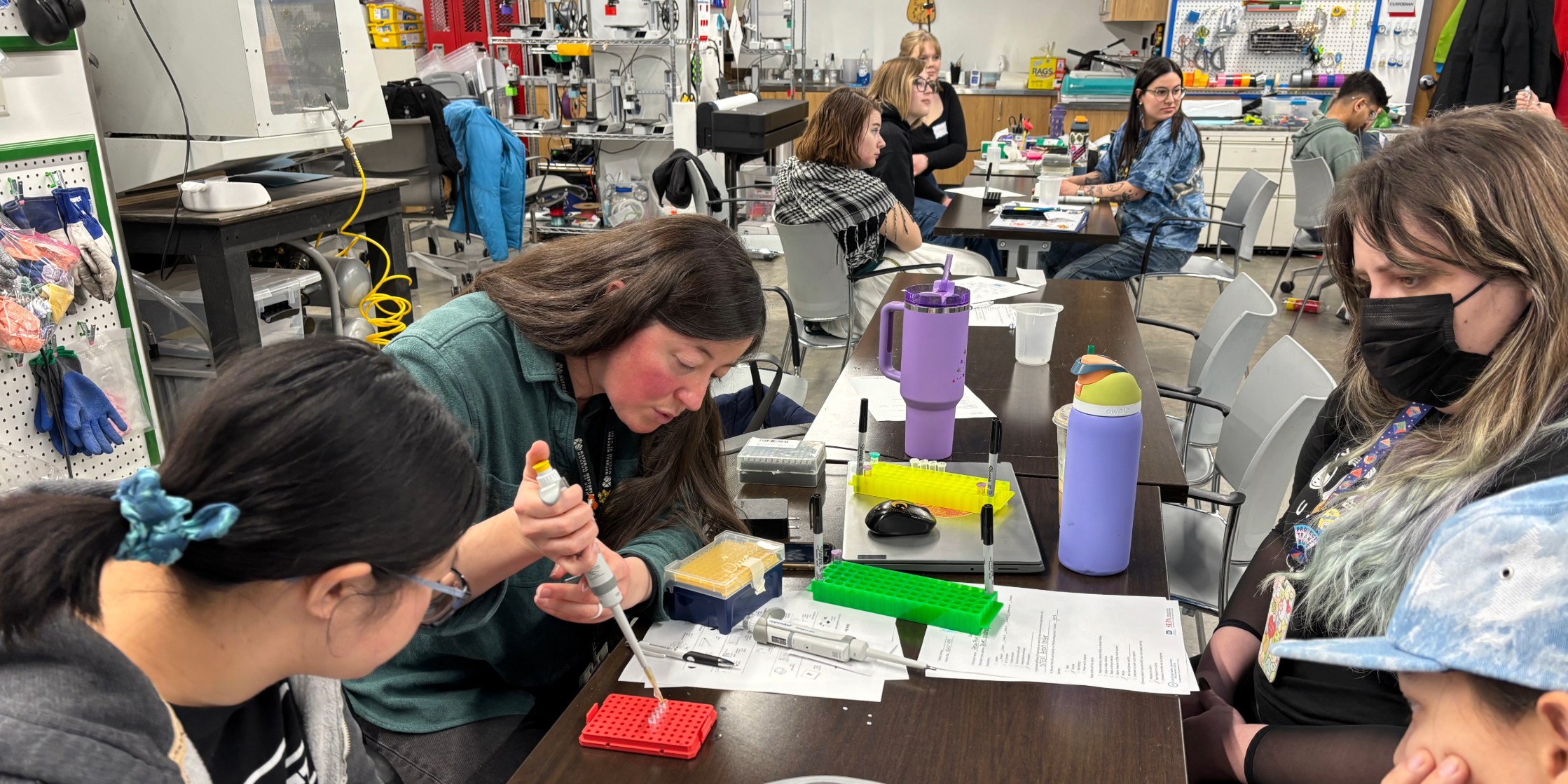 People working with lab equipment at a table in a busy, modern laboratory classroom.
