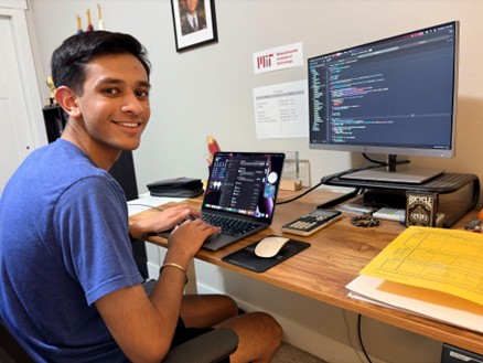 Young man smiling at desk with a laptop and monitor displaying code in a home office.