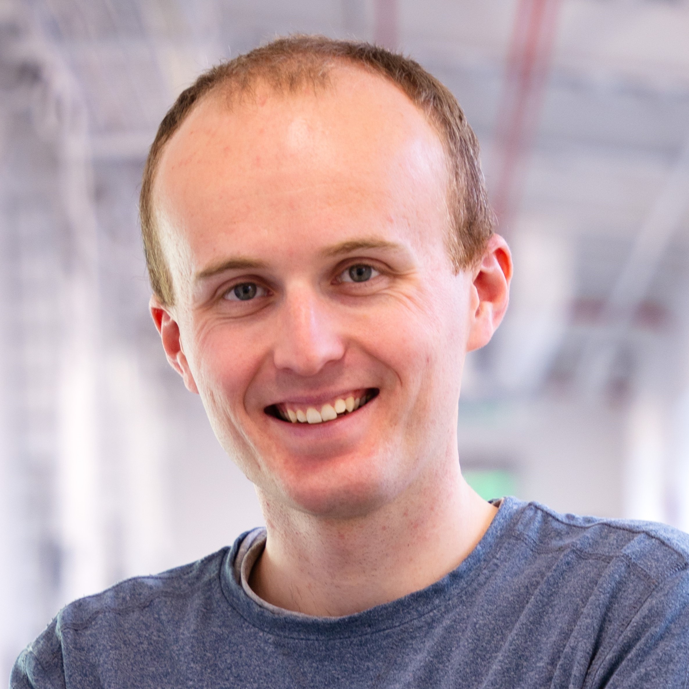A smiling man in a blue shirt stands in a brightly lit indoor hallway.