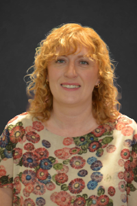 Woman with curly red hair wearing floral top, smiling at the camera against a dark background.
