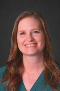 Woman with long brown hair wearing a green top, smiling against a dark gray background.