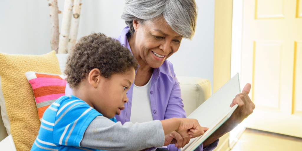 Older woman and young boy sit together on a couch, reading and pointing at a book.