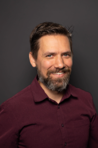 A man with a beard wearing a maroon button-down shirt.