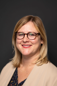 A smiling woman with blonde hair wearing glasses and a blazer.