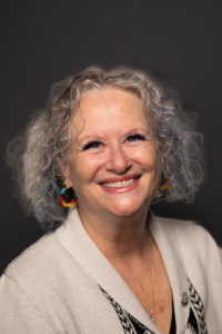Headshot of a smiling woman with wavy gray hair and colorful earrings.